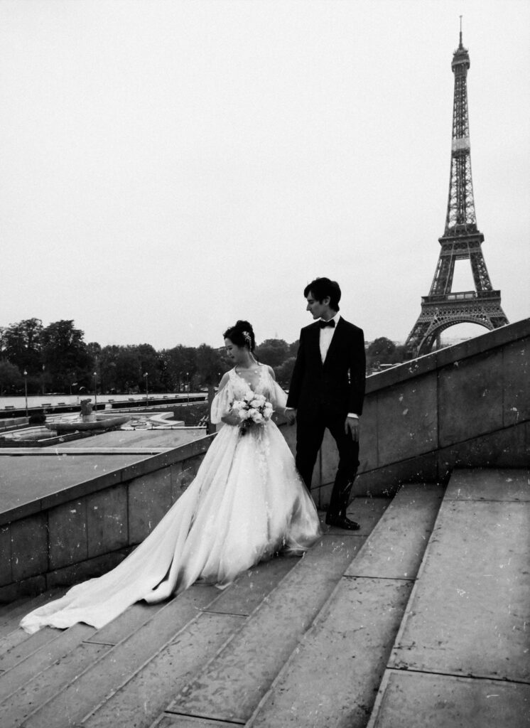 Grayscale Photo of Bride and Groom Standing on Concrete Stairs